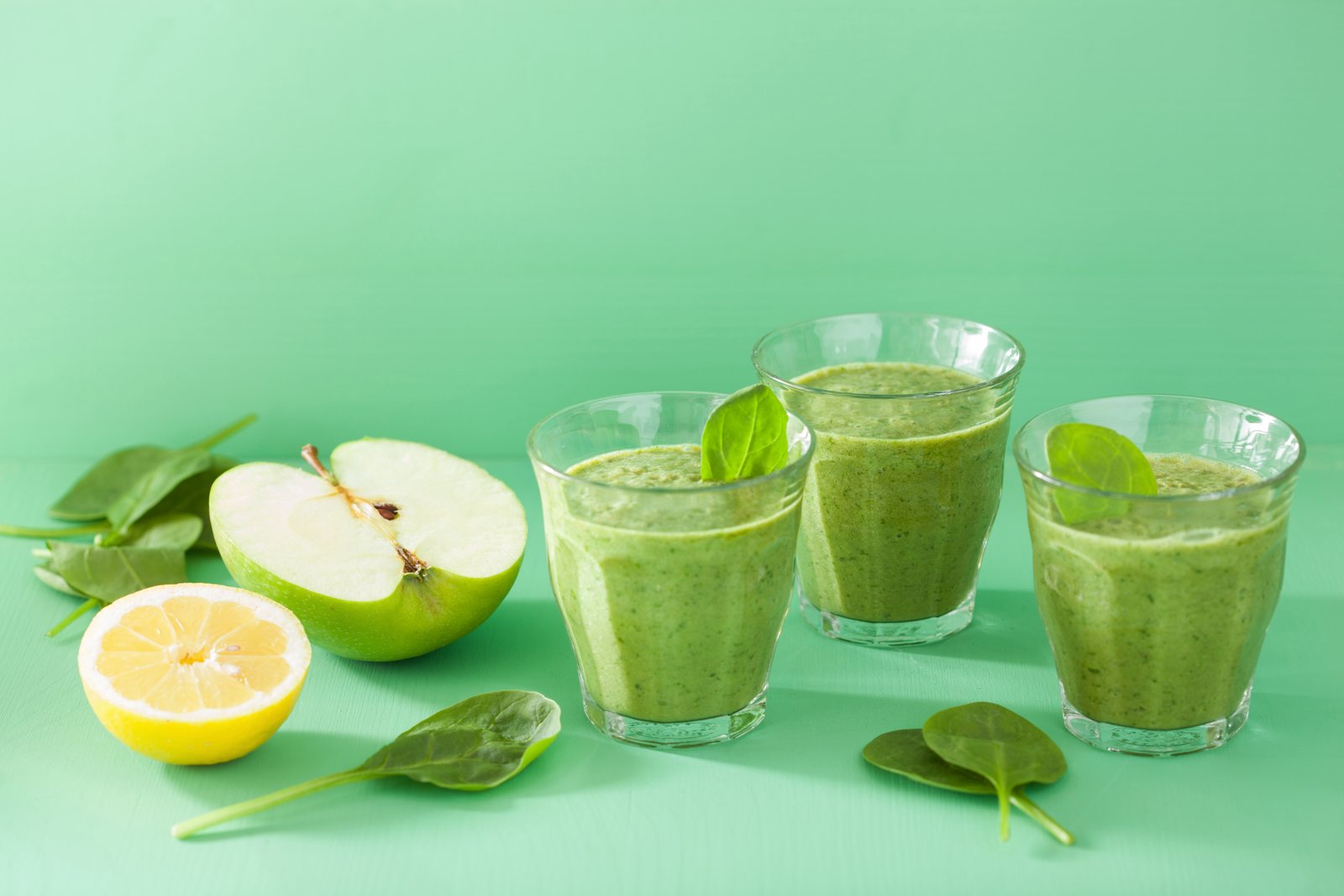 Three green smoothies with apple, lemon, and spinach on a mint-green background