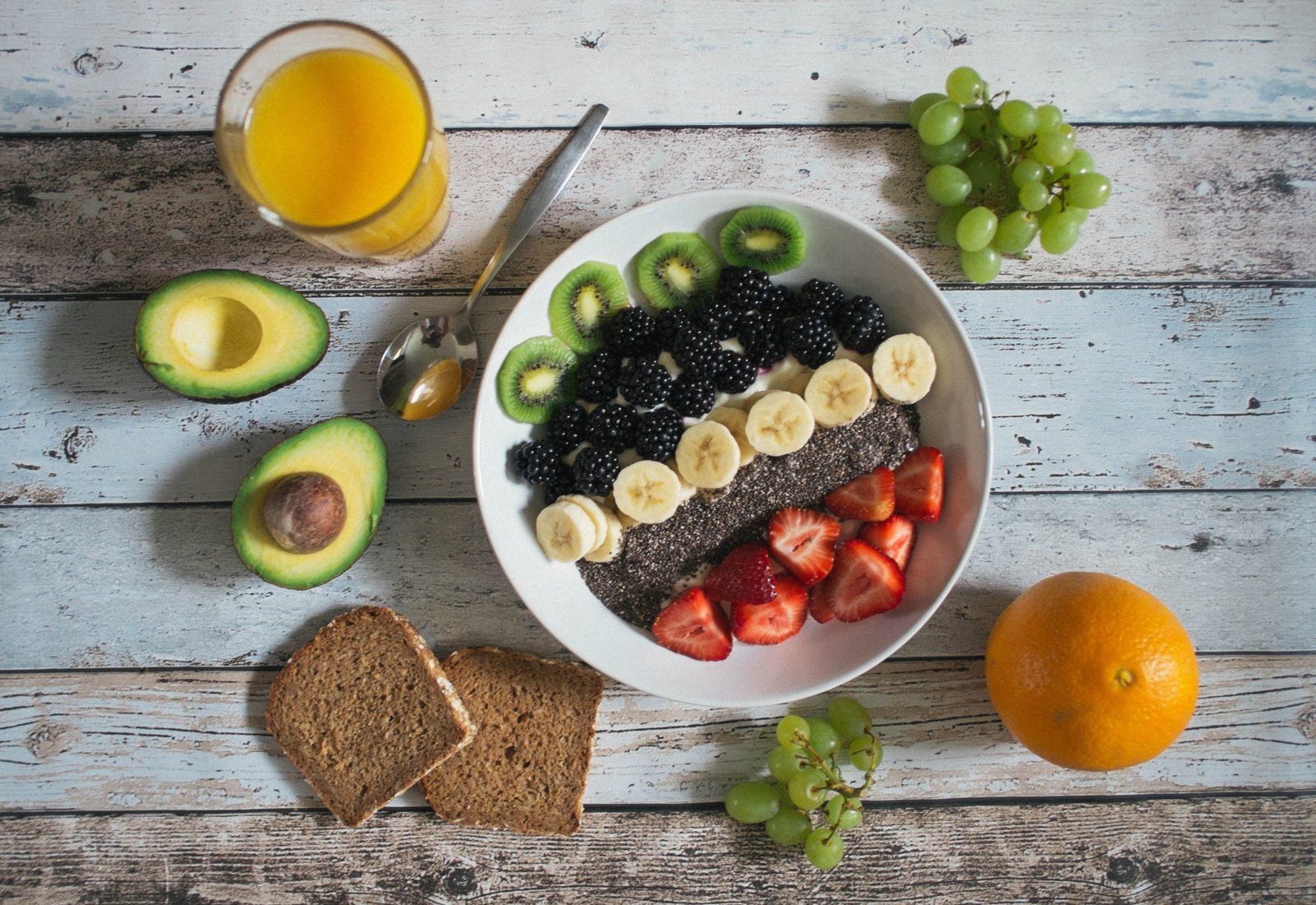 Fruit and yogurt bowl topped with kiwi, blackberries, banana, and strawberries, with orange juice, avocado, grapes, and toast nearby
